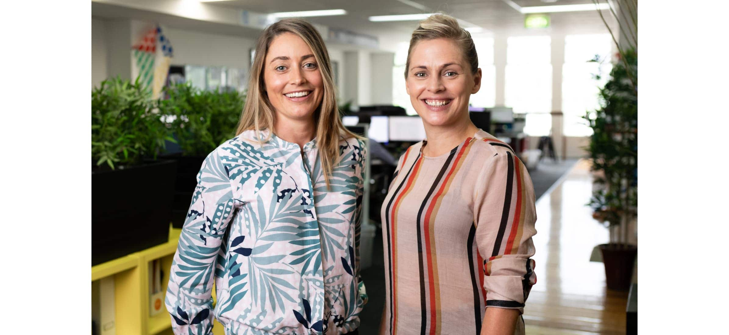 Lucy and Heidi stand by each other smiling at the camera, with their office space behind them.